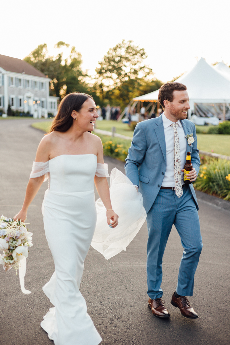 A bride in a white, off-the-shoulder gown walks hand-in-hand with a groom in a light blue suit and floral tie, holding a bottle. They smile outdoors at sunset near the Dennis Inn Wedding venue in Dennis MA.