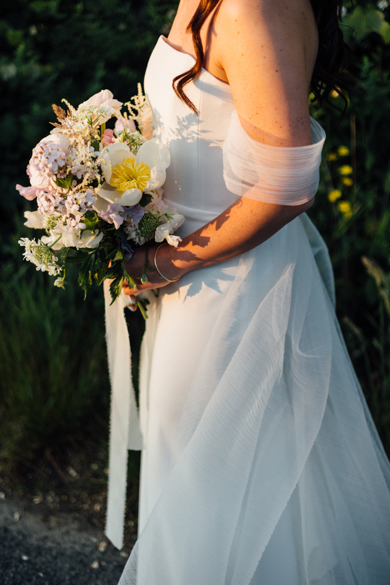 A bride in a strapless white dress holds a pastel bouquet of flowers, standing outdoors in sunlight with greenery at her Dennis Inn Wedding in Dennis, MA. Her hair is loose and wavy, and sheer fabric drapes from her arms.