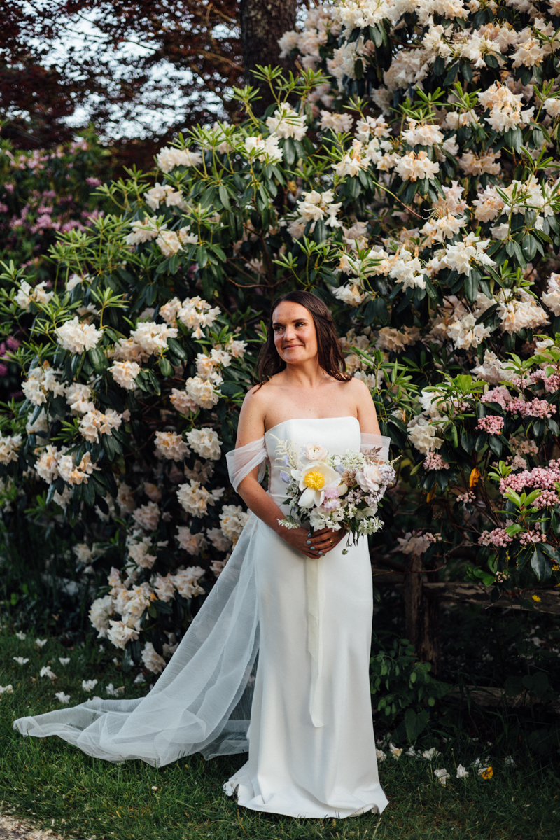A bride in a white, off-the-shoulder gown holds a bouquet and smiles while standing before lush bushes with white and pink flowers at her beautiful Dennis Inn Wedding in Dennis, MA.