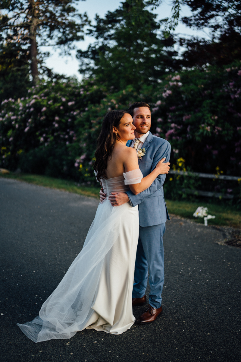 A bride in an off-shoulder white gown and a groom in a blue suit embrace on a paved path, surrounded by greenery and flowering bushes, with soft evening sunlight at their Dennis Inn Wedding in Dennis MA.