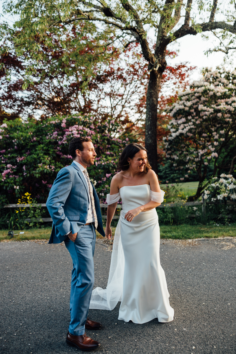 A bride in a white gown and a groom in a blue suit stand together outdoors, smiling and holding hands, surrounded by blooming trees and flowers on a sunny Cape Cod wedding day at the Dennis Inn in Dennis, MA.