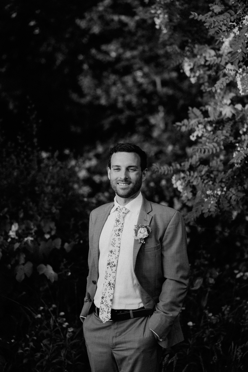 A man in a suit with a floral tie and boutonniere stands outdoors, smiling with his hands in his pockets. Surrounded by leafy foliage, he poses for a timeless black-and-white photo at a Dennis Inn Wedding in Dennis MA.