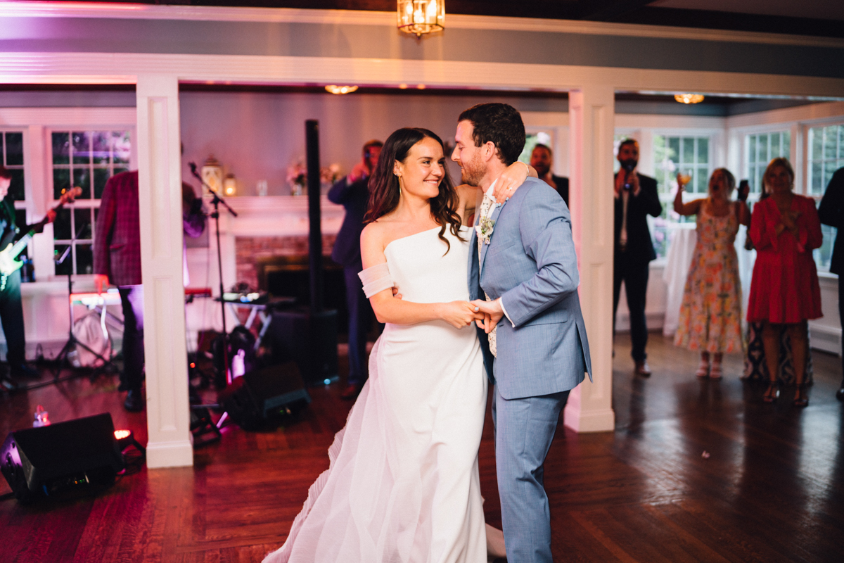 A bride in a white gown and a groom in a light blue suit share their first dance at their Dennis Inn Wedding in Dennis, MA, smiling at each other as guests and a live band watch in the background.