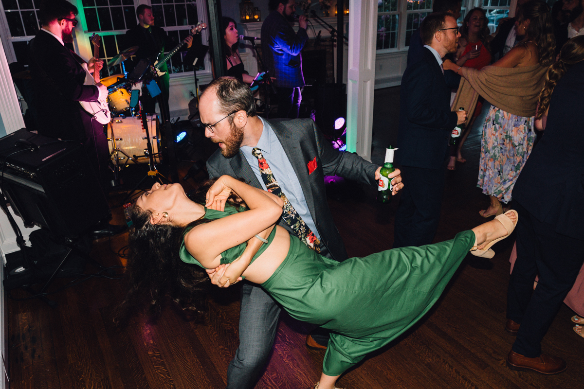 A man in a suit playfully dips a woman in a green dress while dancing at a lively Dennis Inn wedding in Dennis, MA, with a live band performing and other guests socializing in the background.