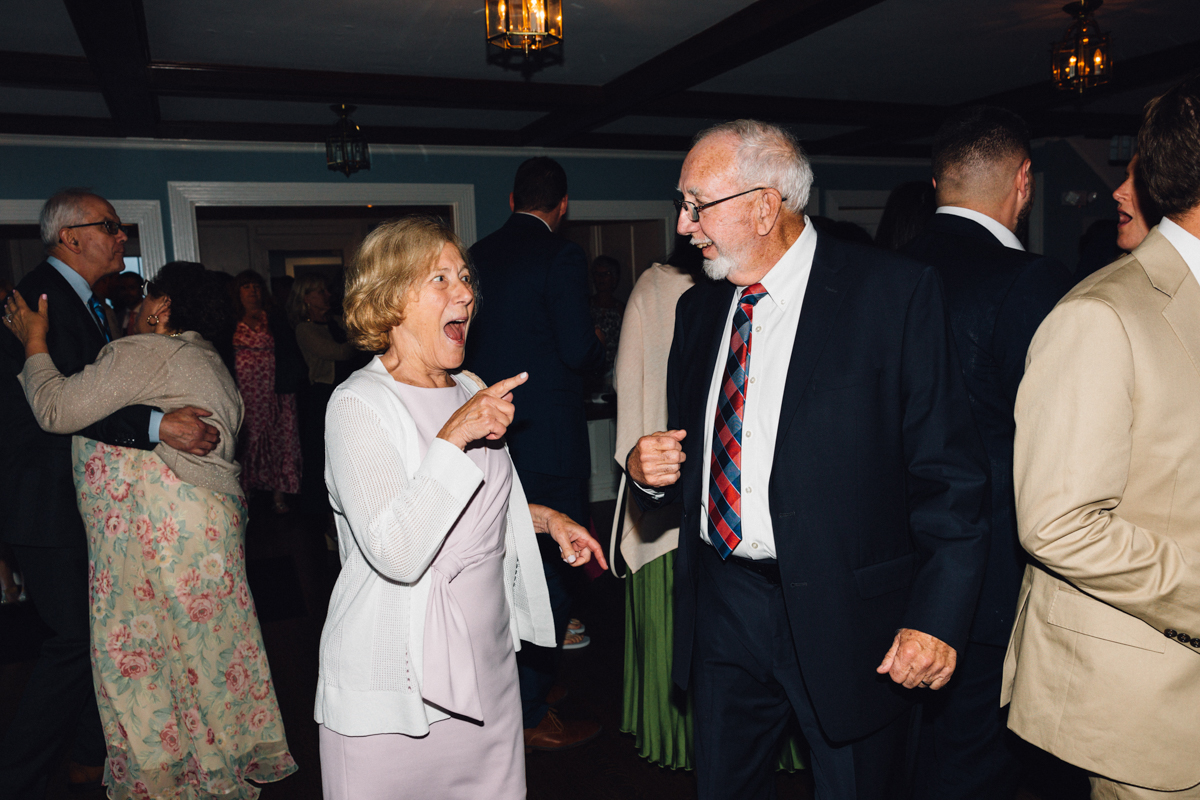 An older woman and man, both dressed formally, smile and dance together at a lively indoor Dennis Inn Wedding, surrounded by other people mingling and dancing in the background.