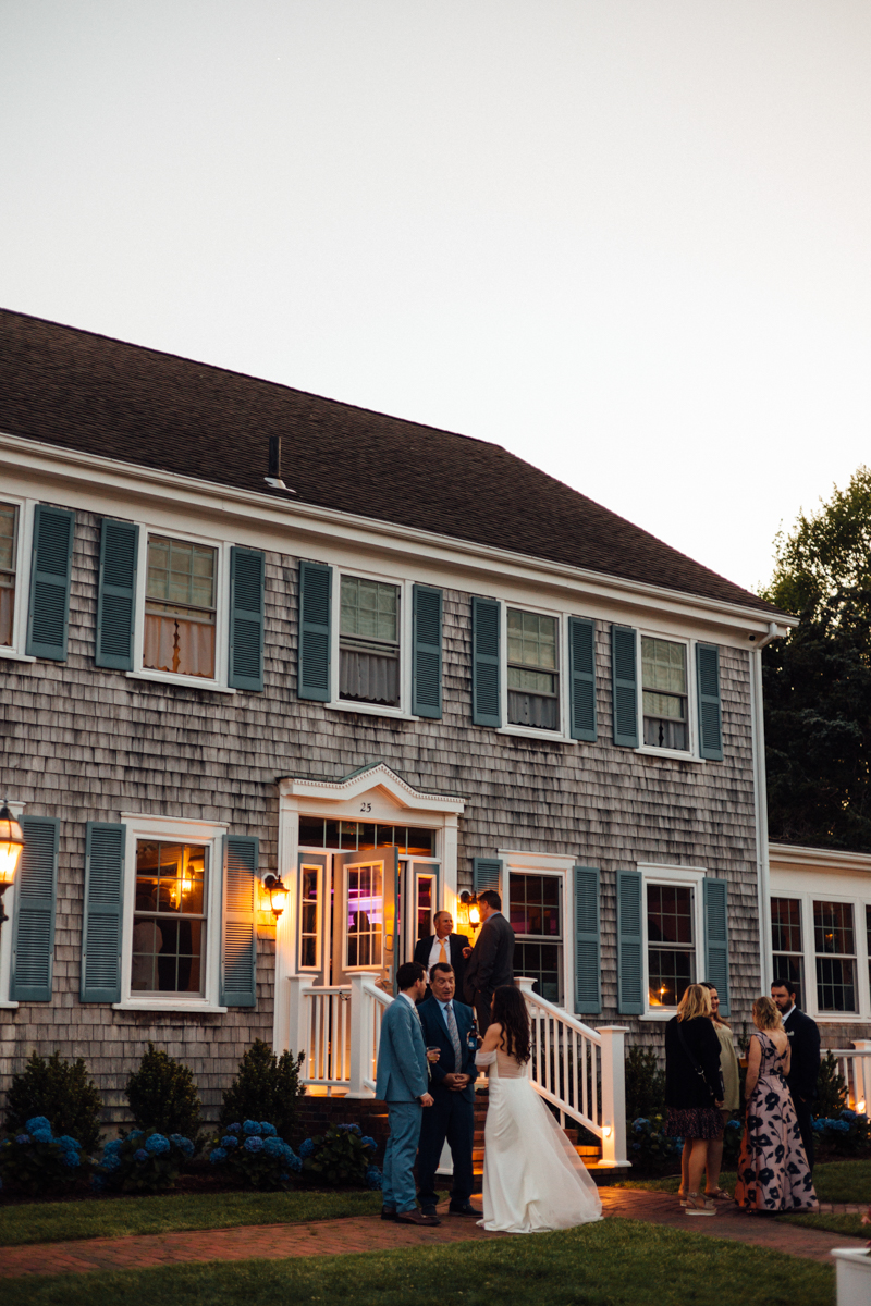 A bride and groom stand with guests outside the Dennis Inn in Dennis, MA—a large grey shingle house with blue shutters at dusk, warm lights glowing from the windows as other guests converse nearby on the lawn.