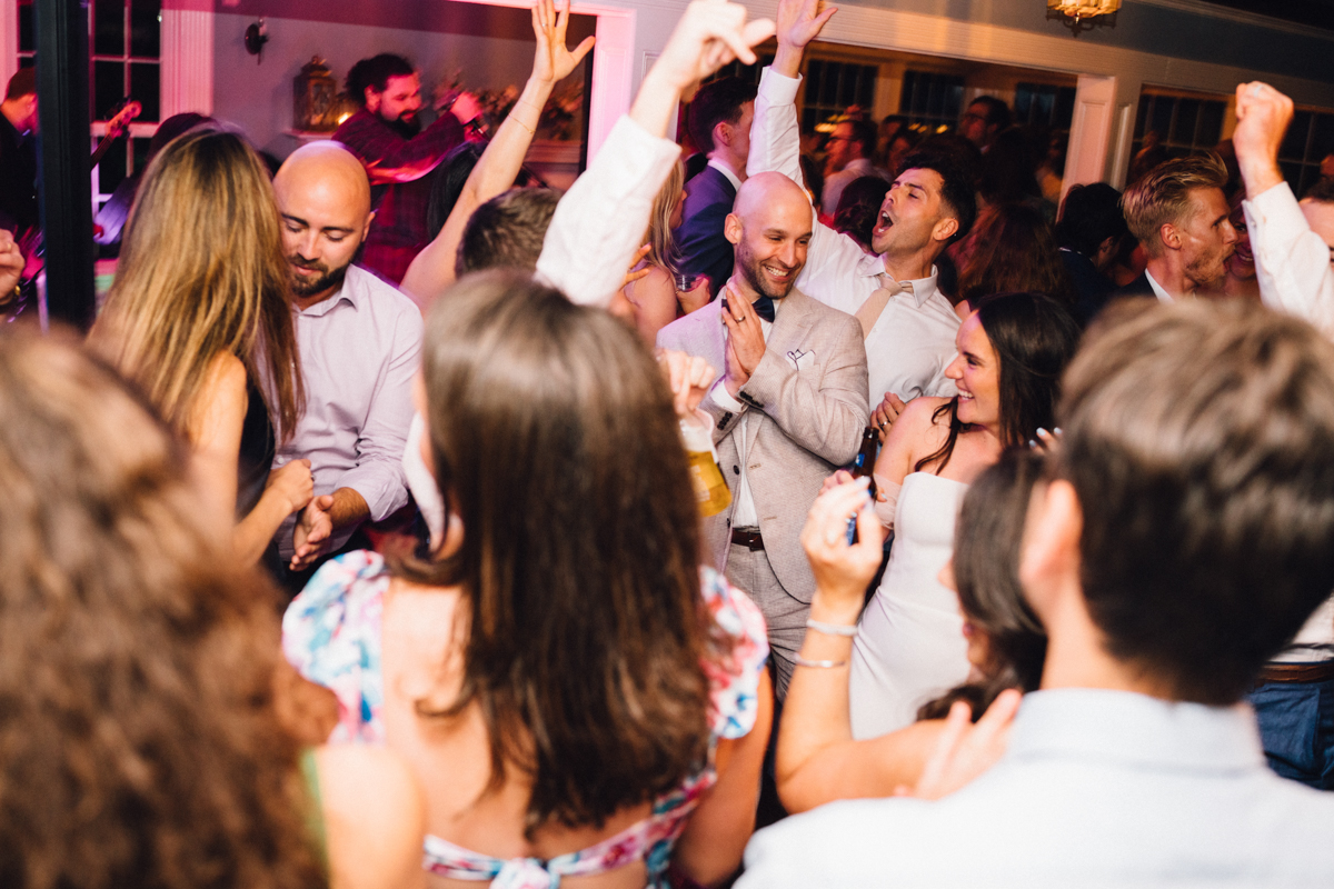 A group of people dressed in semi-formal attire dance and celebrate joyfully at a Dennis Inn Wedding in Dennis MA. Some have their hands raised, laughing and smiling, while a band plays energetically in the background.