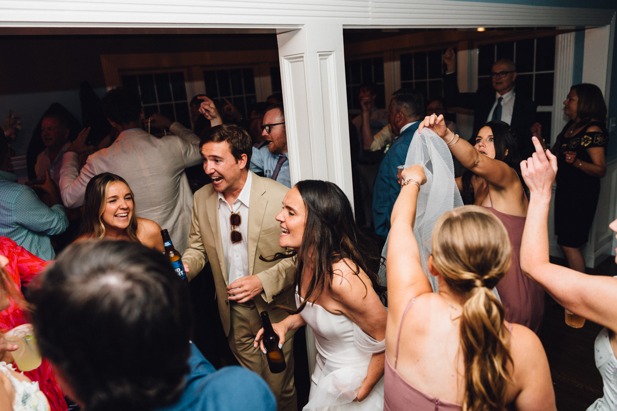 A lively group of people dance and celebrate indoors at a Dennis Inn Wedding. Some are holding drinks, and a woman in a white dress lifts a veil, smiling among friends. The atmosphere is joyful and energetic in Dennis, MA.