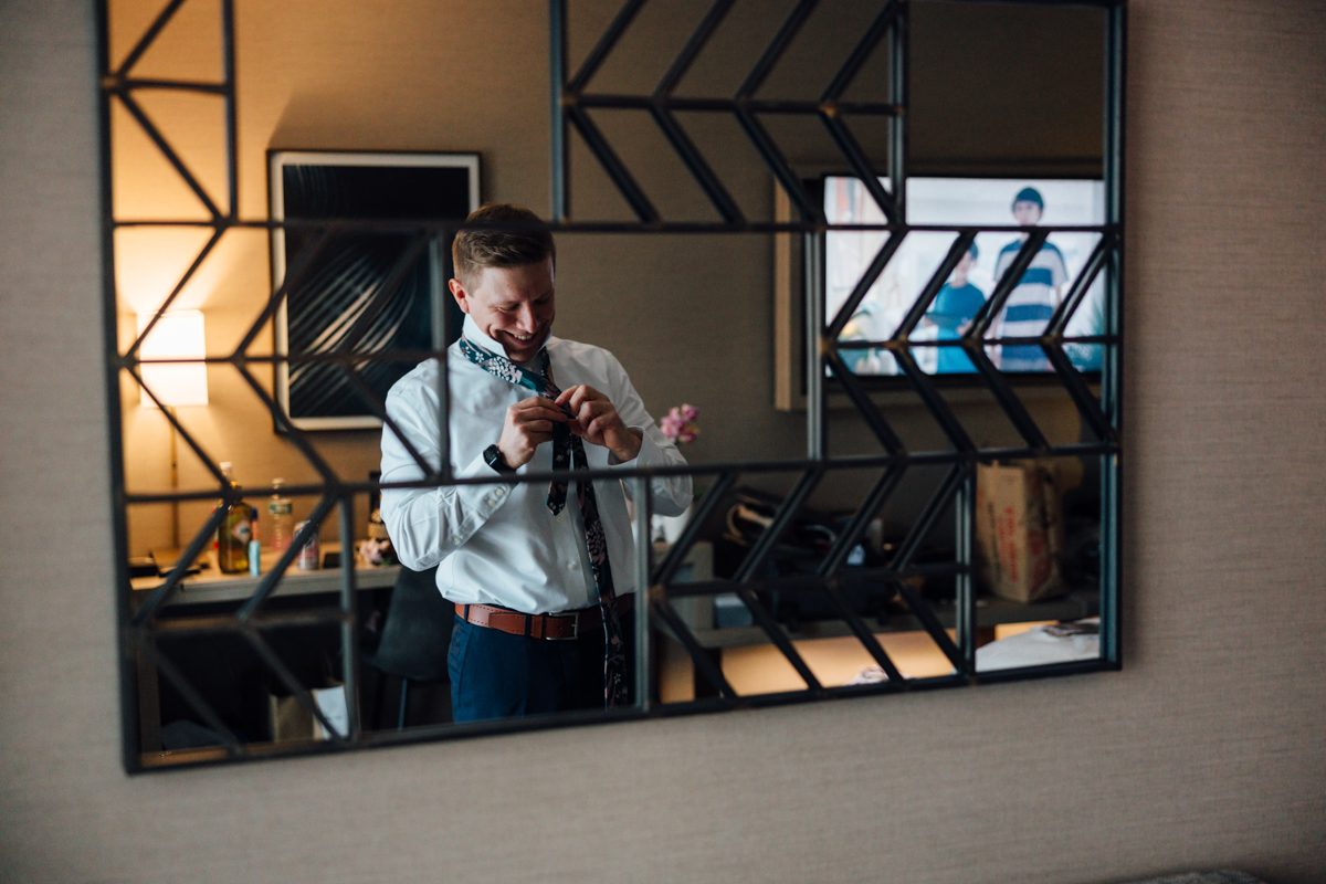 A man, reflected in a geometric wall mirror, smiles while tying his tie in a modern room with a desk, lamp, TV, and various items in the background.