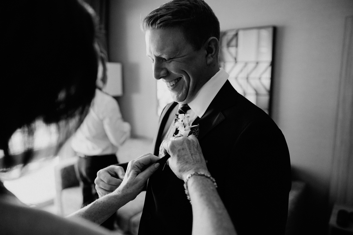 A smiling man in a suit has a boutonniere pinned to his lapel by another person. The scene is indoors, and the moment appears candid and joyful. The photo is black and white.