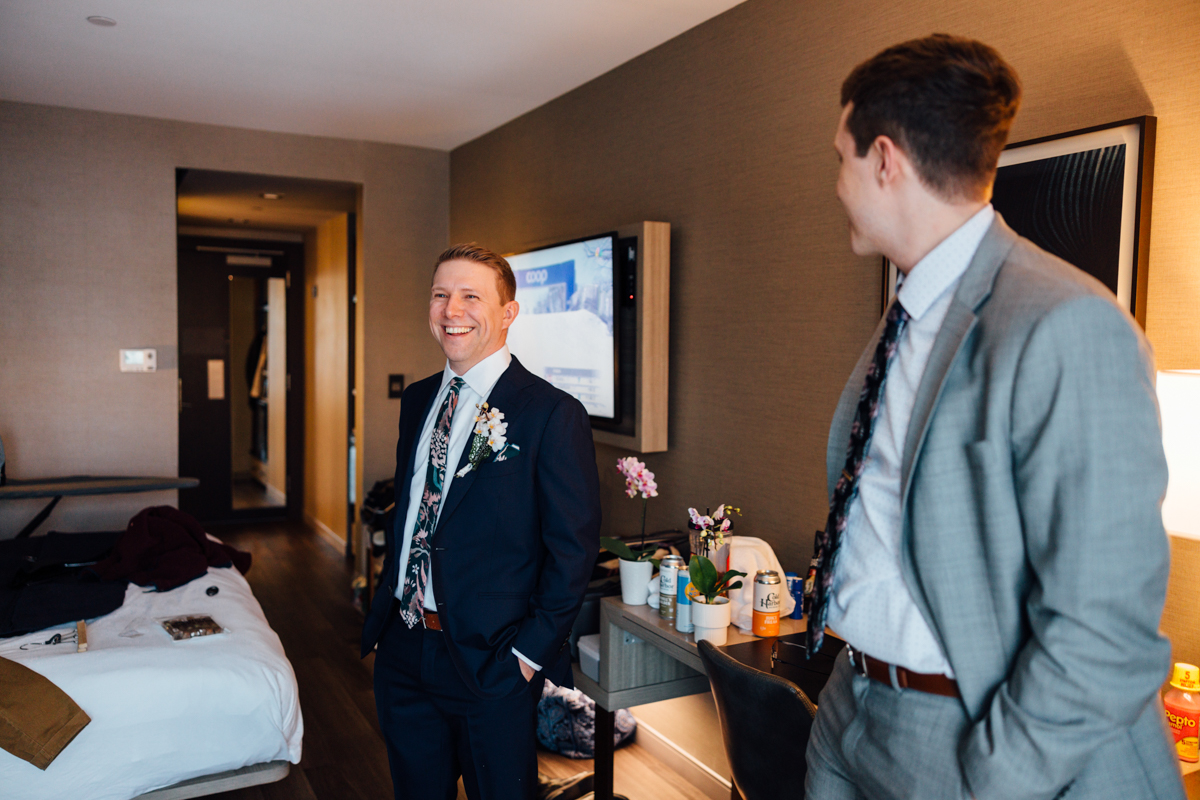 Two men in suits stand and smile in a modern hotel room; one faces forward, wearing a boutonniere, while the other looks at him. The room has a bed, desk, TV, and some drinks on the table.