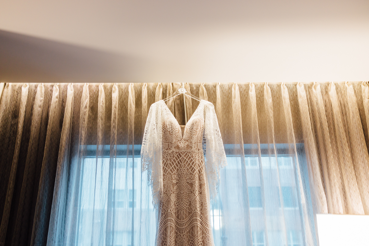 A lacy white wedding dress hangs on a hanger in front of a window with sheer curtains, with soft natural light illuminating the room.