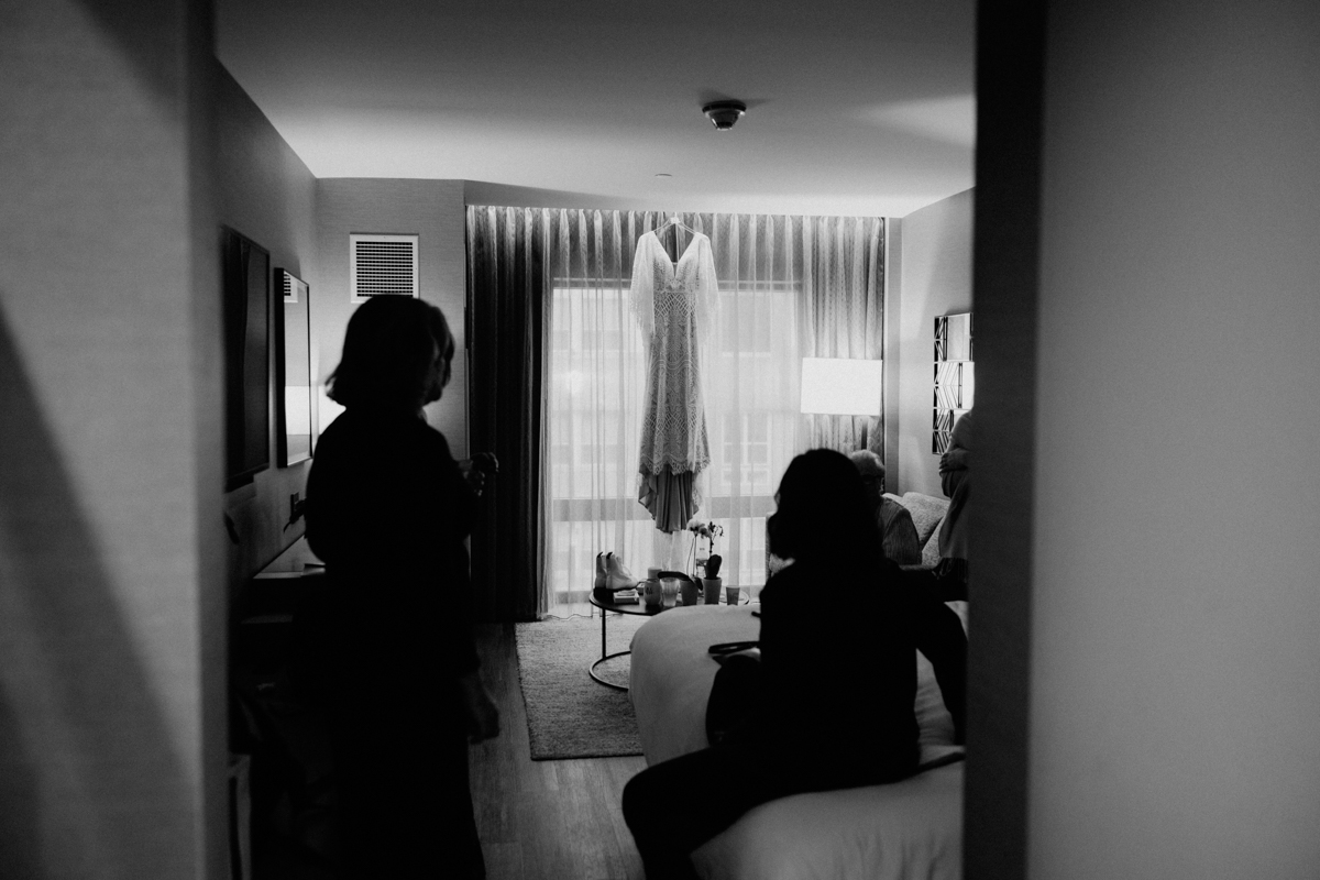 A black and white photo of a hotel room with a wedding dress hanging in front of a window. Captured by a New England Botanic Garden Tower Hill Wedding photographer, two silhouetted people are in the foreground, one on the bed, one near the doorway.