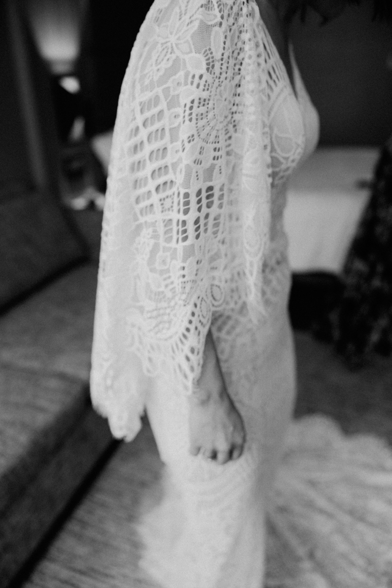 A black and white photo showing a person in a detailed lace dress with flowing sleeves, standing indoors. The focus is on the lace pattern while the background and face are blurred.