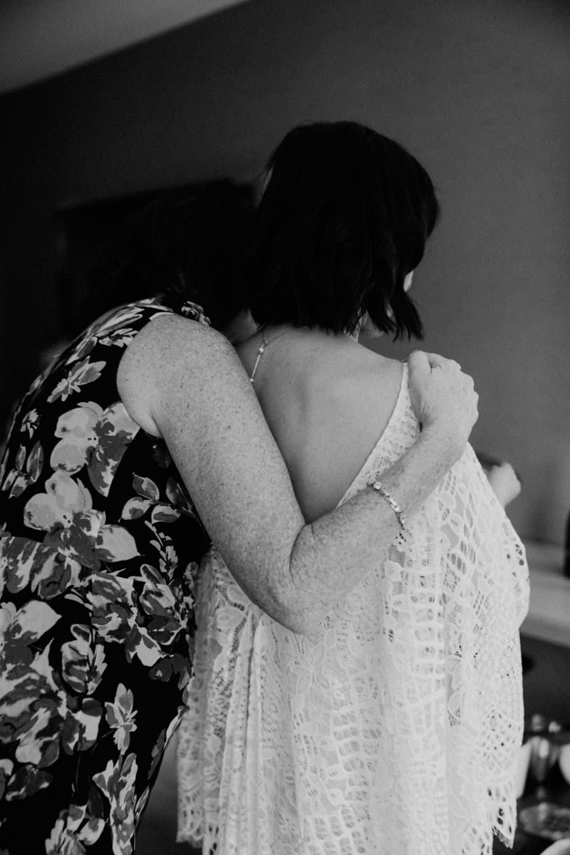 Two women stand close together with their backs to the camera; one in a floral dress wraps her arm around the other, who wears a white lace garment. The image is in black and white, conveying a tender, intimate moment.