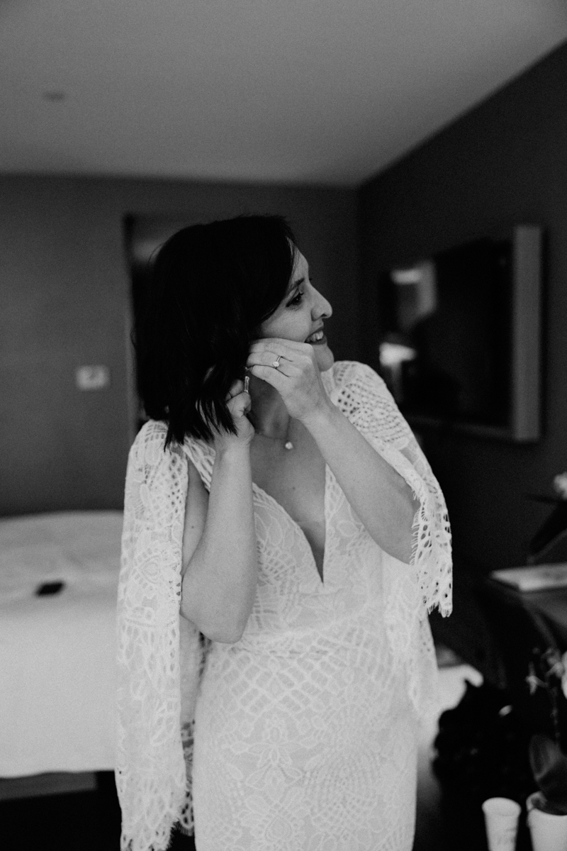 A woman in a white lace dress adjusts her earring and smiles, standing in a softly lit room with a bed and a television in the background. The photo is in black and white.