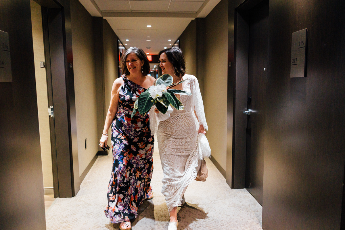 Two women walk side by side in a hallway lined with doors. One wears a floral dress and the other, in a white lace gown, holds a bouquet and a white shawl. Both are smiling and appear happy.