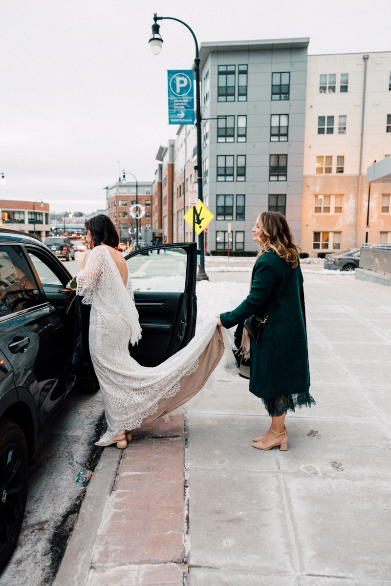 A bride in a white dress steps into a car while another woman in a green coat holds her train on a snowy city street—captured by New England Botanic Garden Tower Hill wedding photographer, with tall buildings and parked cars in the background.