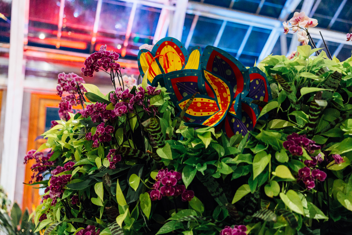 A colorful, decorative flower sculpture is nestled among lush green leaves and clusters of purple flowers inside a greenhouse with a glass ceiling.