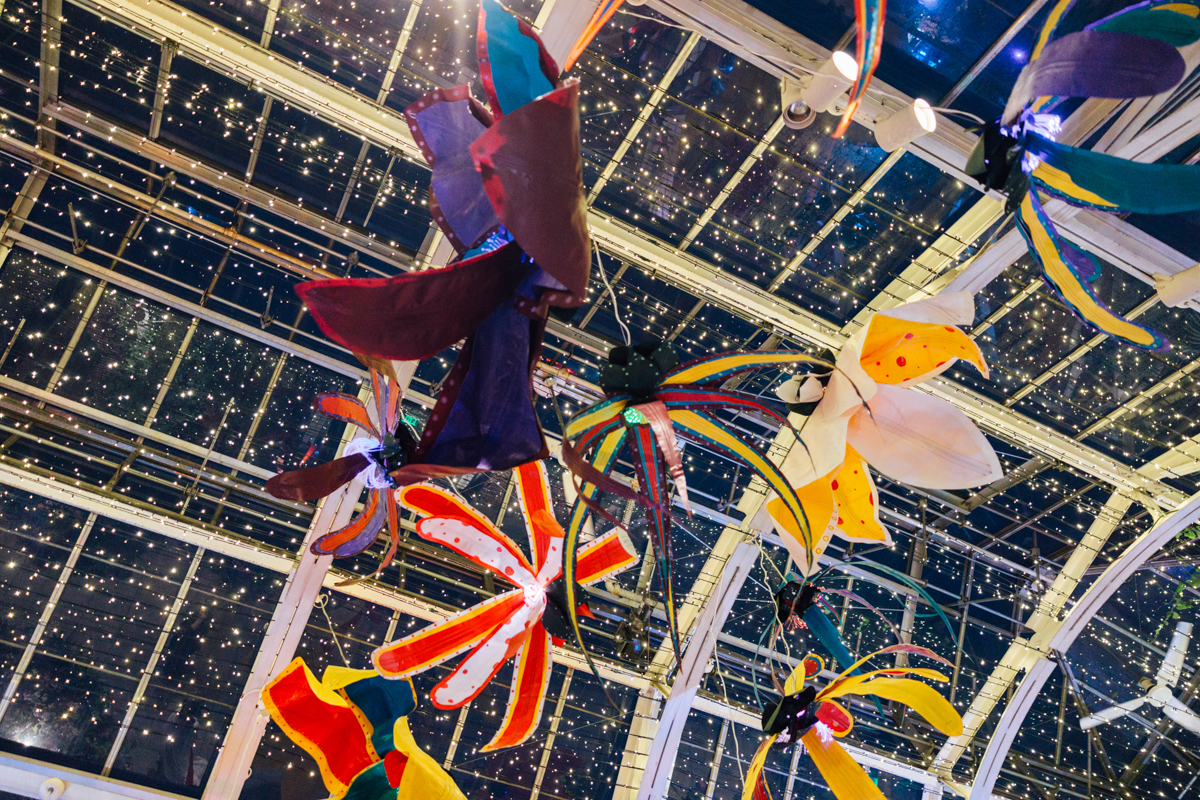 Colorful, flower-shaped decorations hang from the ceiling of a glass-roofed structure lit with string lights, creating a whimsical, festive atmosphere against a starry night-like backdrop.