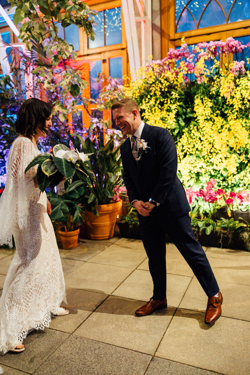 A bride in a white lace dress and a groom in a navy suit smile at each other in a greenhouse filled with vibrant flowers and lush greenery.