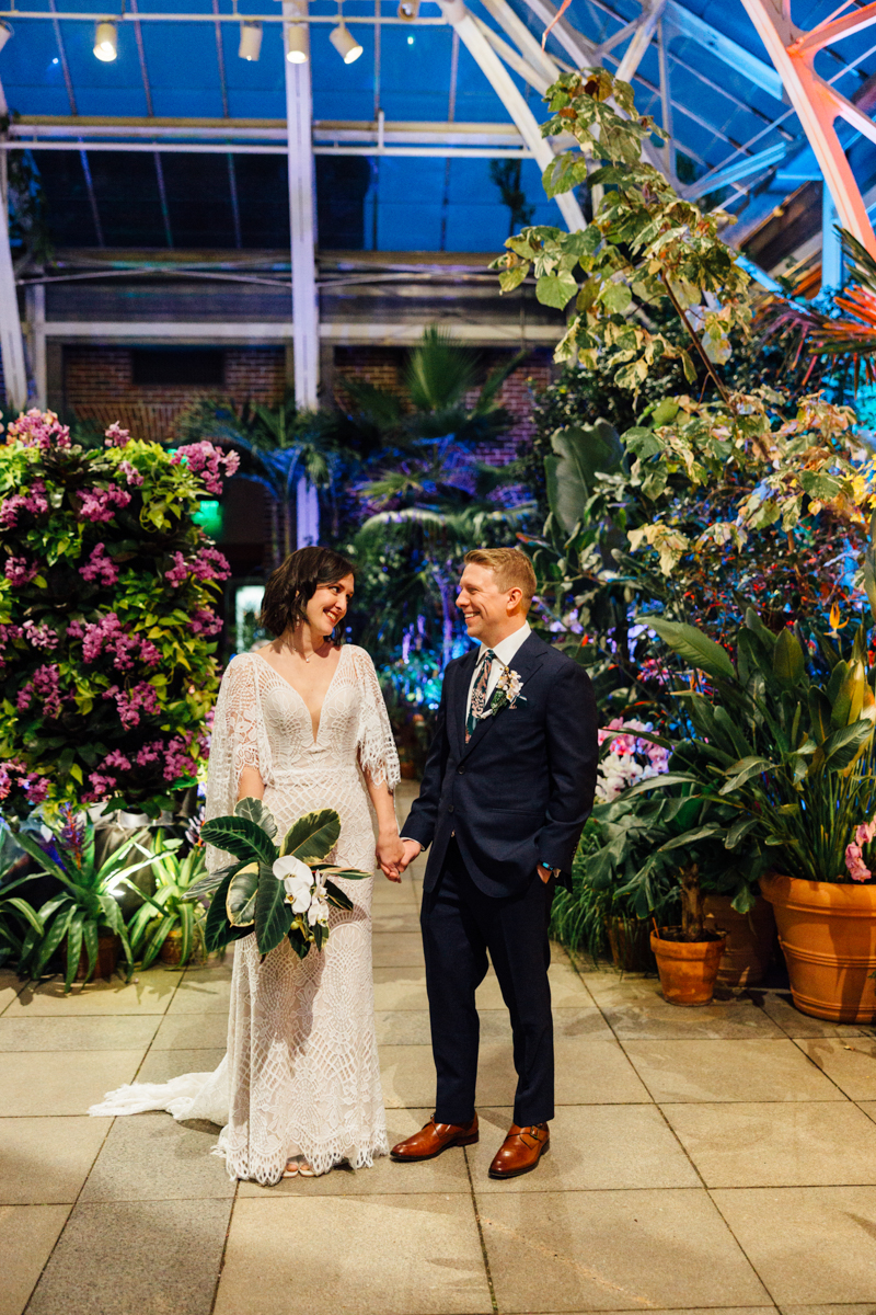 A bride in a lace dress and groom in a dark suit stand holding hands and smiling at each other in a lush, indoor botanical garden with vibrant green plants and purple flowers.