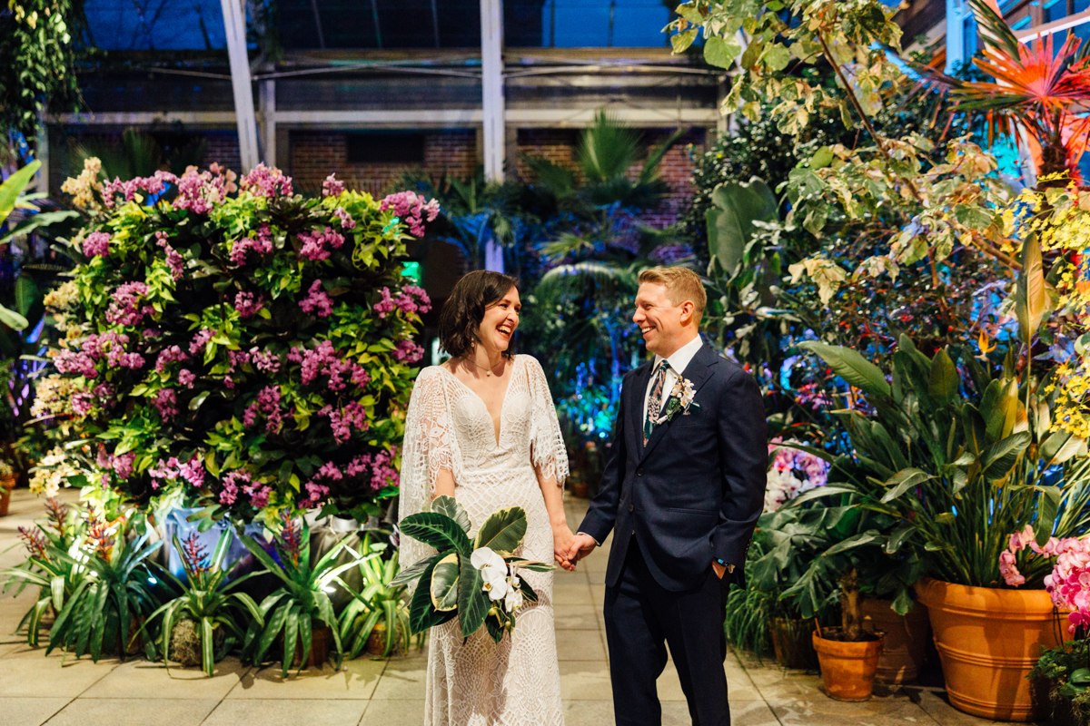 A bride and groom hold hands and smile at each other in the lush indoor garden of New England Botanic Garden Tower Hill, surrounded by colorful flowers and greenery. Captured by a talented wedding photographer, their joy shines brightly.