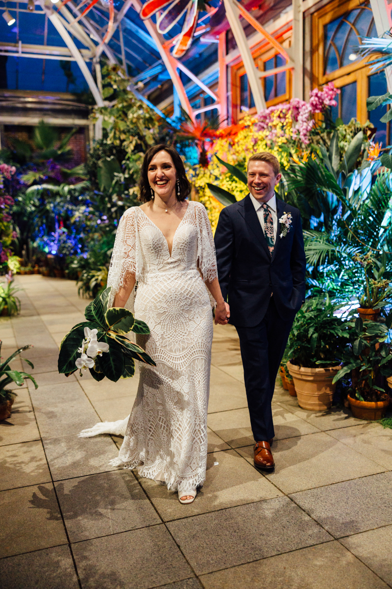 A bride in a white lace dress holds a bouquet and walks hand-in-hand with a groom in a dark suit and floral tie inside a lush, colorful greenhouse filled with vibrant plants and flowers. Both are smiling happily.