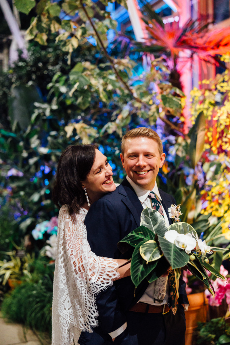 A joyful couple dressed in wedding attire stands in a lush, colorful indoor garden. The bride hugs the groom from behind, both smiling, with tropical plants and vibrant lighting in the background.