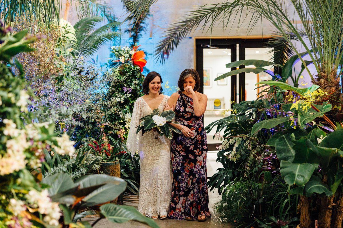 A smiling bride in a white lace dress walks down a lush, tropical aisle at New England Botanic Garden Tower Hill, with a woman in a floral gown wiping away tears—captured perfectly by an attentive wedding photographer amid vibrant plants and colorful flowers.