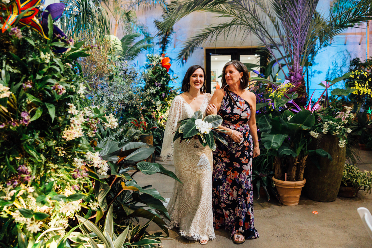A bride in a white lace dress walks with a smiling woman in a floral gown through a lush indoor garden filled with tropical plants and colorful flowers. Both look happy and elegant.