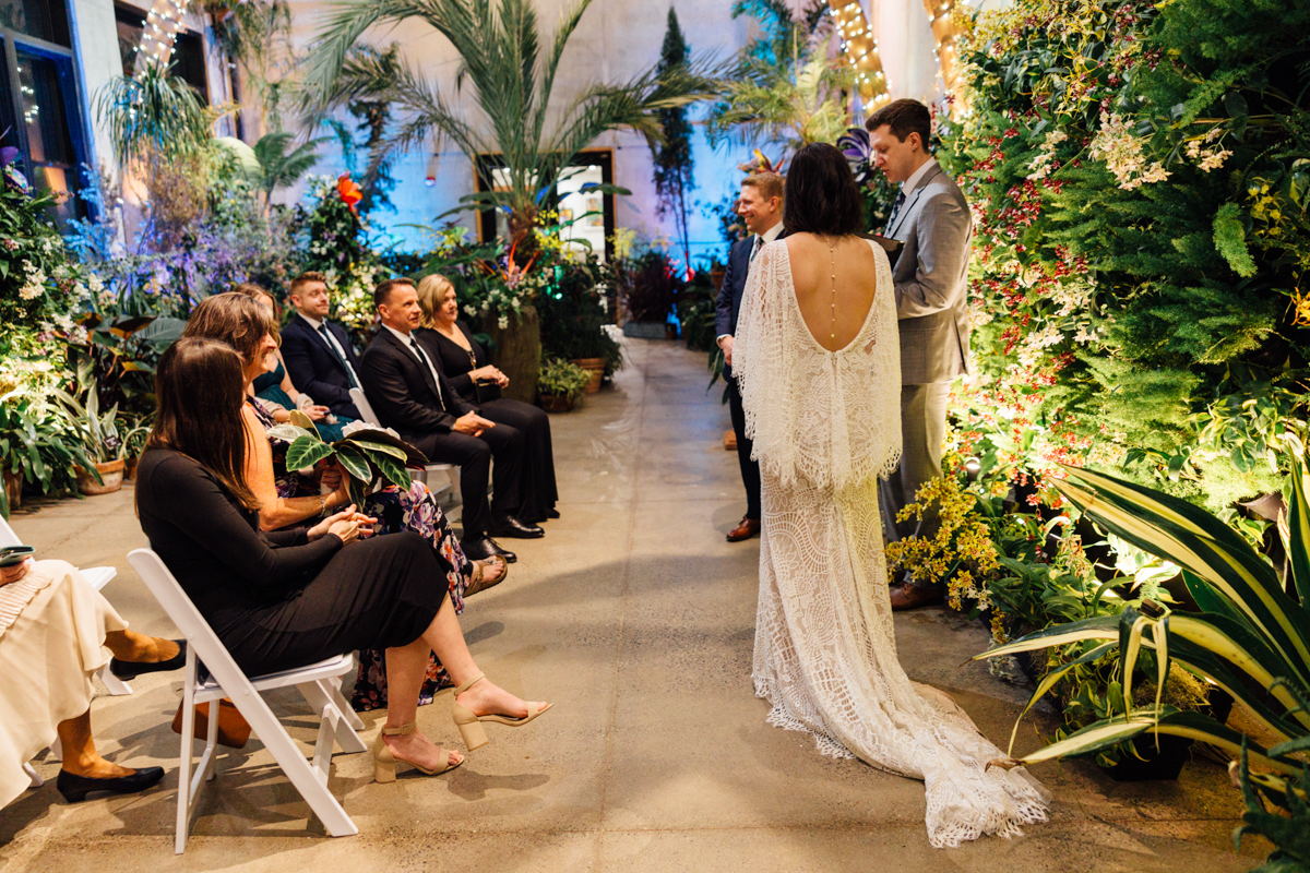 A bride in a long, lacy white dress stands with a groom at an indoor garden wedding ceremony, surrounded by lush green plants and seated guests watching attentively.