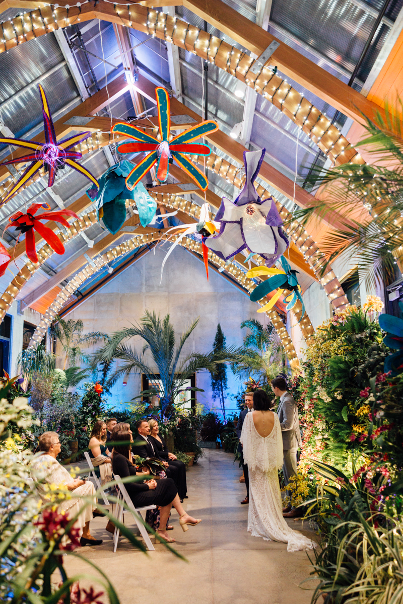 A couple stands at the altar during their wedding ceremony in a vibrant, plant-filled venue with colorful, oversized flower decorations and string lights hanging from the arched ceiling. Guests are seated on both sides.