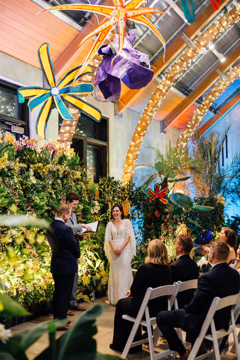 A bride and groom stand at the altar during an indoor wedding ceremony, surrounded by guests. The space is decorated with lush greenery, colorful oversized flowers, and string lights on arched wooden beams.