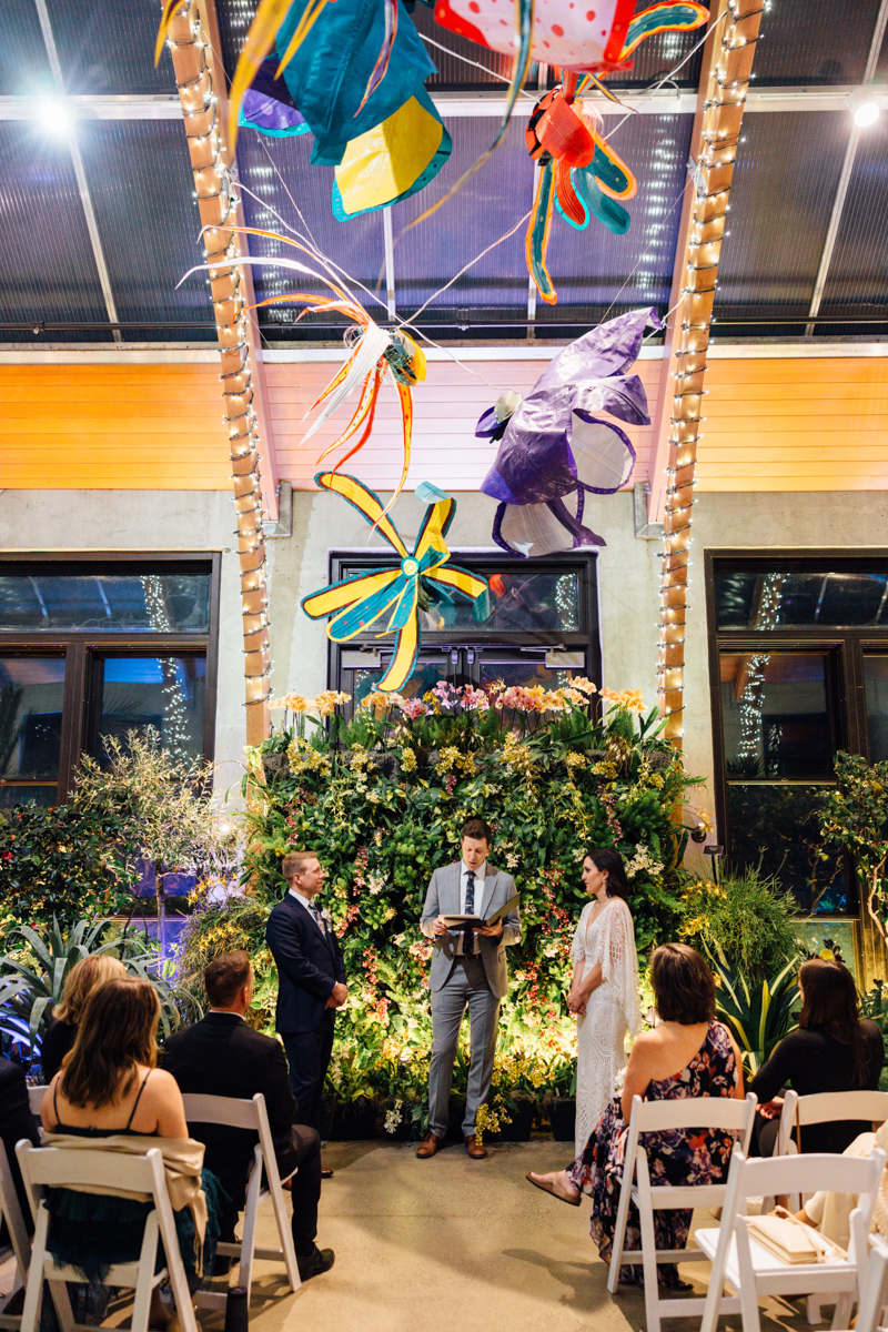 A couple stands facing each other at their indoor New England Botanic Garden Tower Hill wedding ceremony, with a lush floral backdrop and an officiant between them, as guests sit in white chairs beneath colorful, oversized insect sculptures.