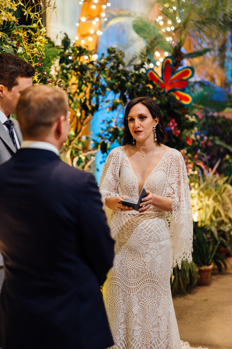A bride in a white lace dress reads vows from a booklet during a wedding ceremony, standing in front of two people in suits, surrounded by colorful flowers and lush greenery.