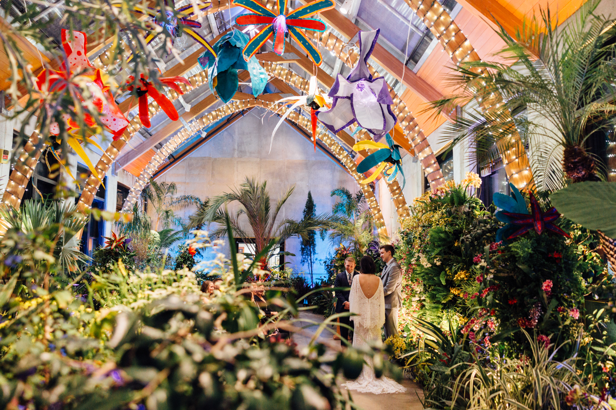 A couple stands under brightly lit arches adorned with colorful, oversized flowers in the lush indoor tropical garden at New England Botanic Garden Tower Hill, captured perfectly by a wedding photographer amidst green plants and festive hanging lights.