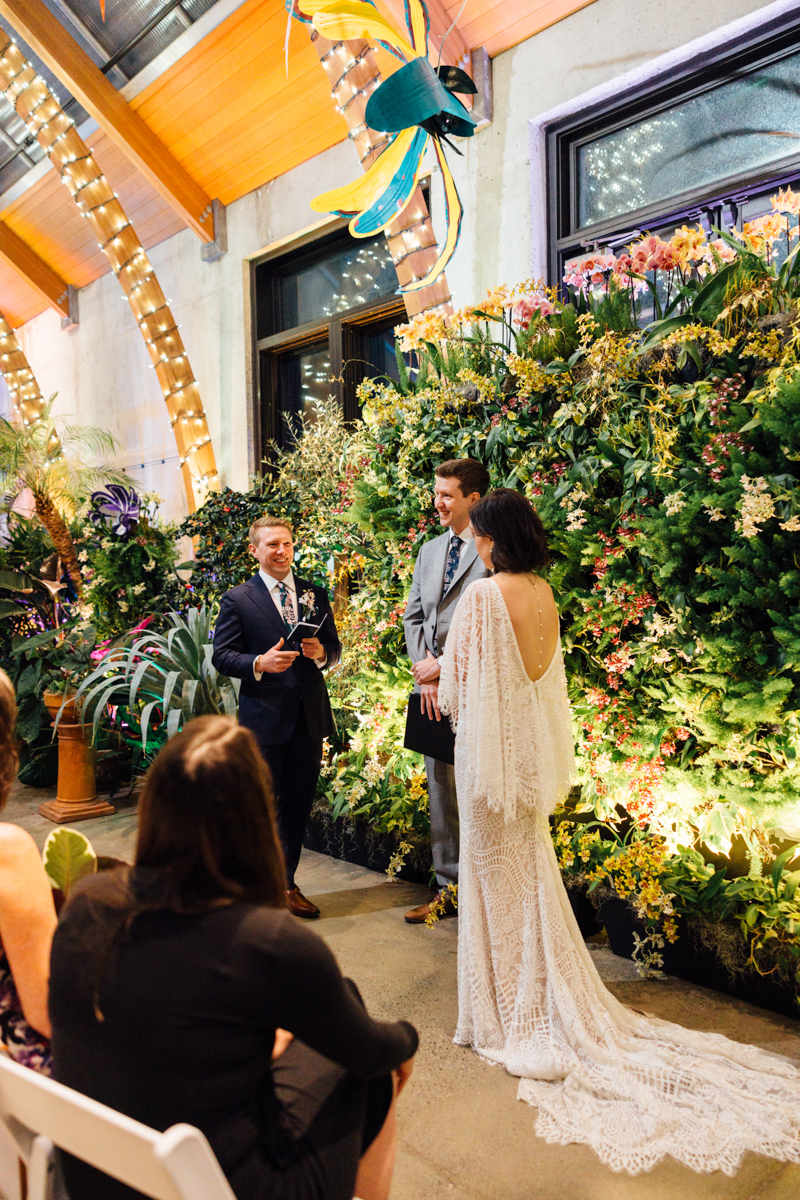 A bride and groom stand facing each other during their indoor wedding ceremony, with an officiant speaking. They are surrounded by lush greenery and flowers, with guests seated nearby under warm, decorative lighting.