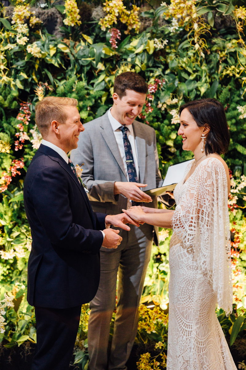 A bride and groom exchange rings during their wedding ceremony, smiling at each other. The officiant stands behind them, holding a book, with lush greenery and flowers in the background.