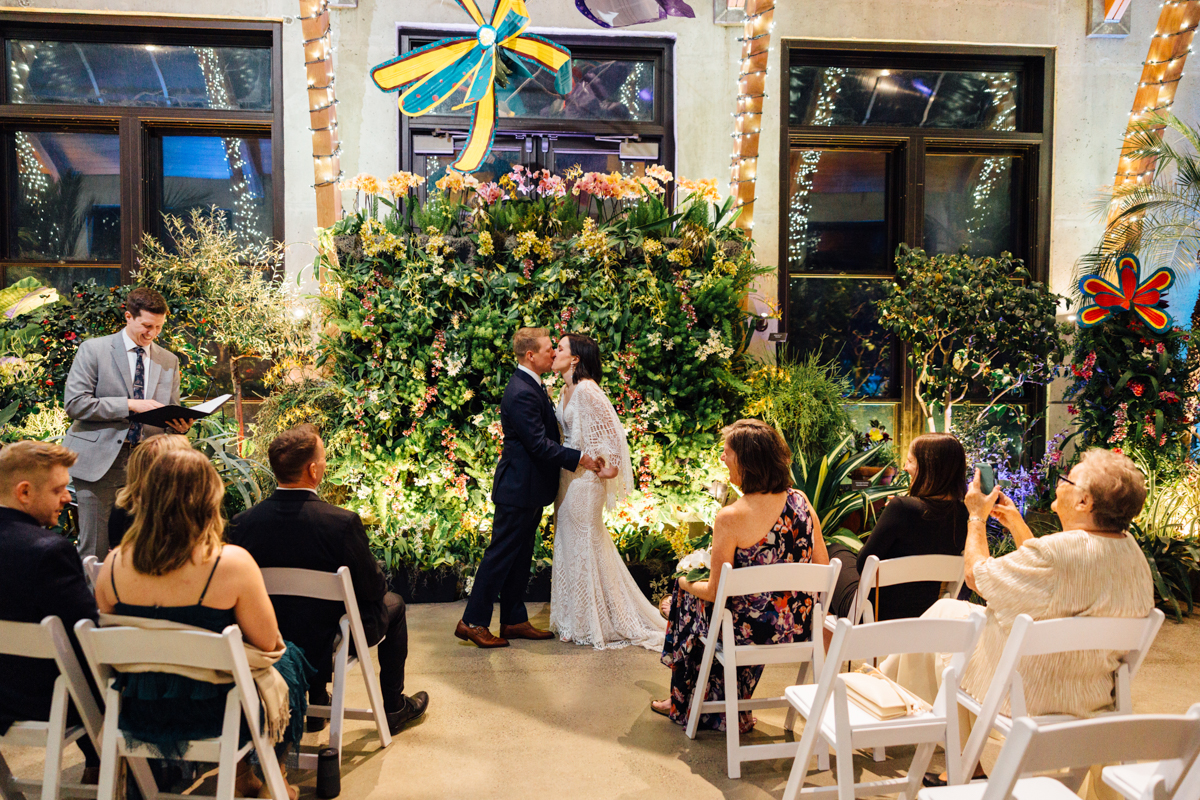 A bride and groom share a kiss at their indoor wedding ceremony, surrounded by lush greenery and flowers. Guests, seated on white chairs, watch and take photos, while an officiant stands to the side reading from a book.