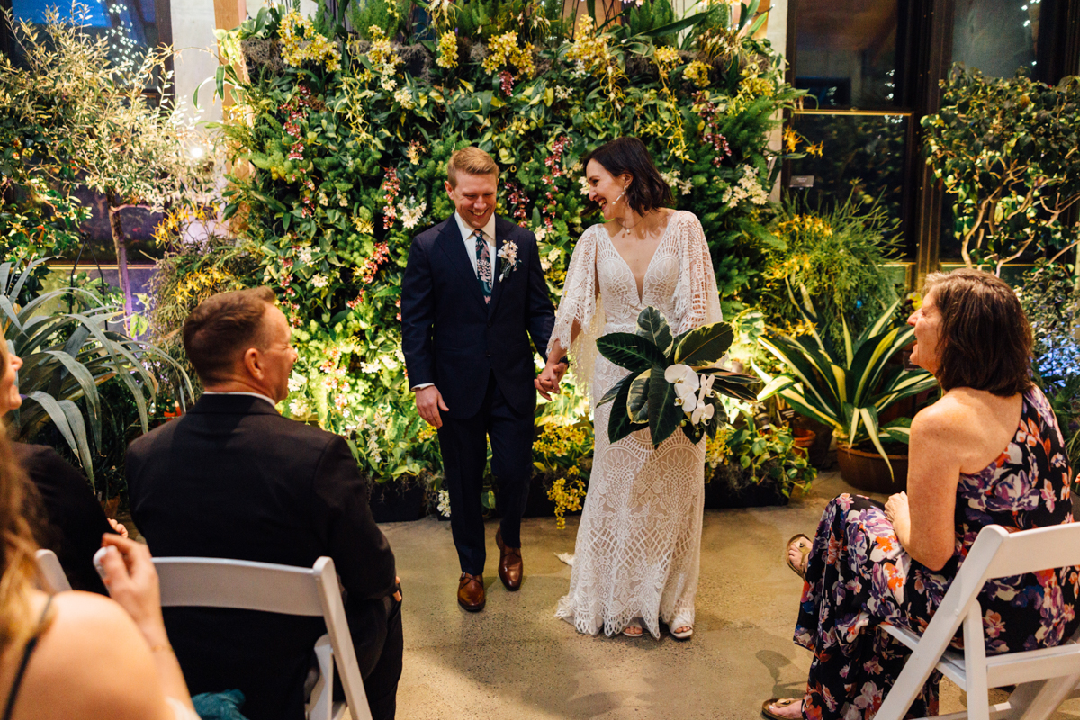 A bride and groom smile as they walk hand in hand down the aisle, surrounded by lush greenery and seated guests at an indoor wedding ceremony. The bride wears a lace dress and holds a bouquet; the groom is in a dark suit.
