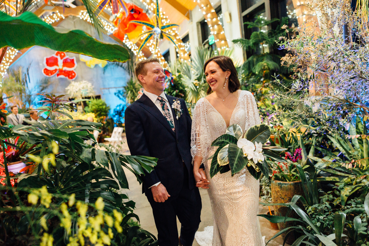 A bride and groom smile at each other while walking hand in hand through a lush, plant-filled indoor space decorated with lights and tropical flowers. The bride holds a bouquet; both look joyful and elegant.
