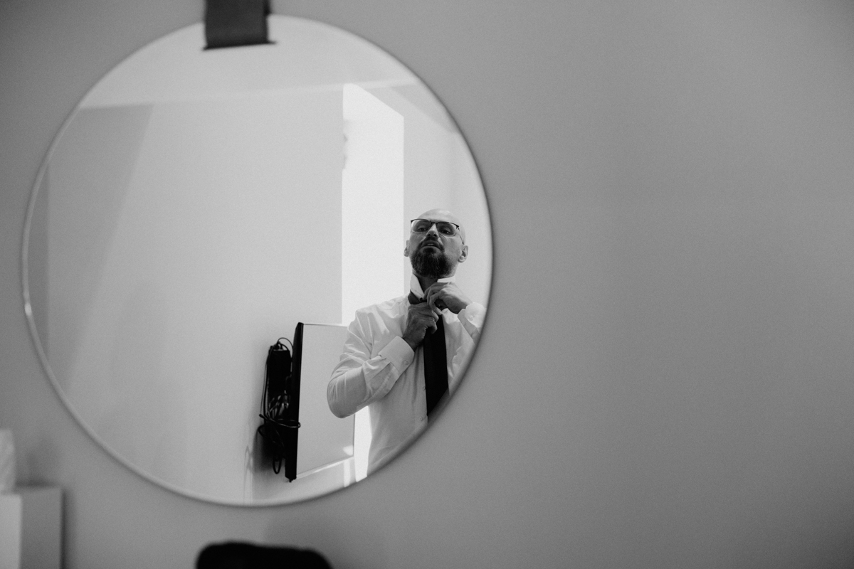 A man adjusting his tie while looking in a round mirror, dressed in a white shirt and black tie in a modern, minimalistic room.