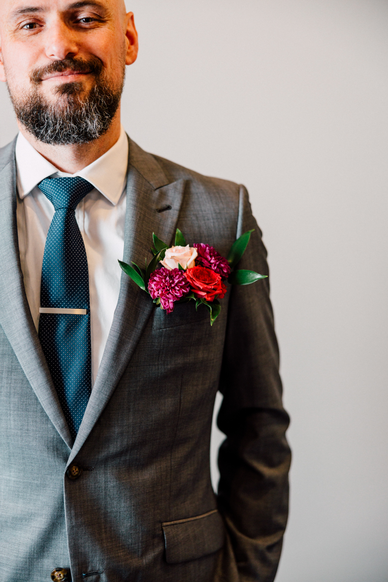 A close-up of a well-dressed man in a gray suit with a polka dot tie, showcasing a vibrant floral boutonniere featuring roses and other blooms.