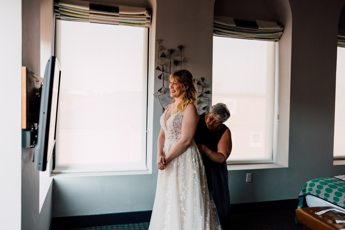 A bride smiles as she is assisted by a woman in fastening her wedding dress in a bright, elegantly decorated hotel room.