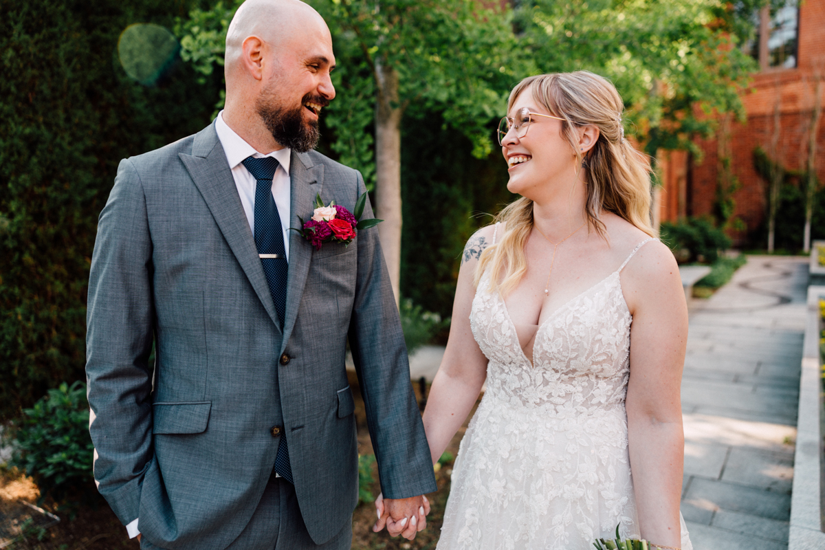 A groom in a gray suit and a bride in a lace dress smile at each other while holding hands in a garden setting.