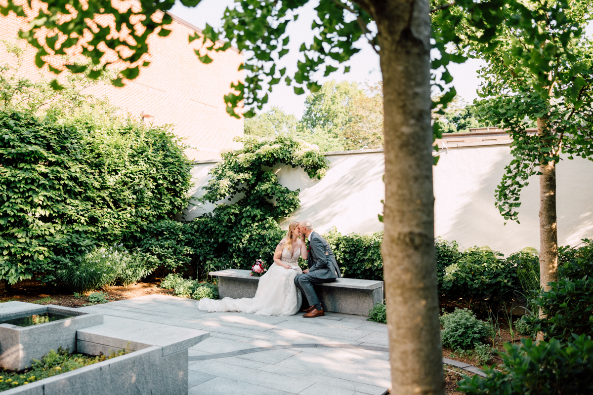 A couple sharing a tender kiss on a stone bench surrounded by lush greenery in a serene garden setting.