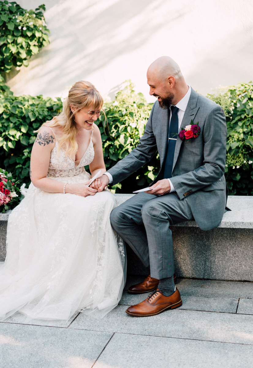 A joyful couple sharing a tender moment, seated on a stone bench surrounded by greenery, with the bride in a lace gown and the groom in a gray suit.