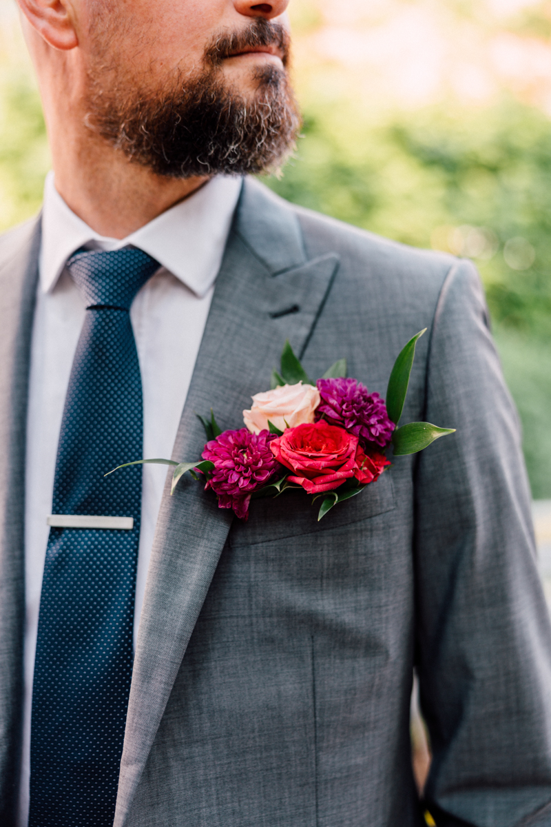 A close-up of a man in a gray suit with a navy tie, showcasing a colorful floral boutonniere featuring roses and other blooms.