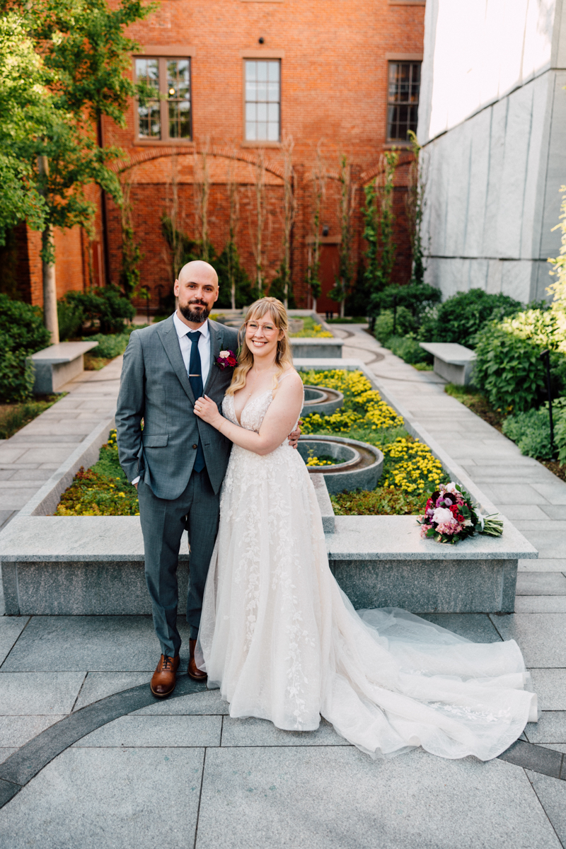 A joyful couple stands together in a beautifully landscaped garden, surrounded by flowering plants and greenery, with the bride in a lace gown and the groom in a gray suit.